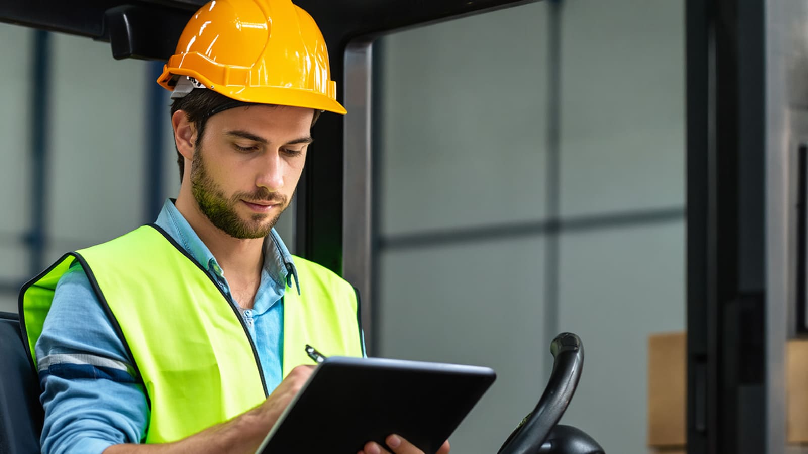 Mitarbeiter mit Schutzhelm und Sicherheitsweste trägt Daten auf einem Tablet in einem Lager oder bei der Arbeit mit einem Gabelstapler ein.